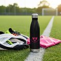 Black soccer mom water bottle with pink logo on a soccer field with shoes and a towel.