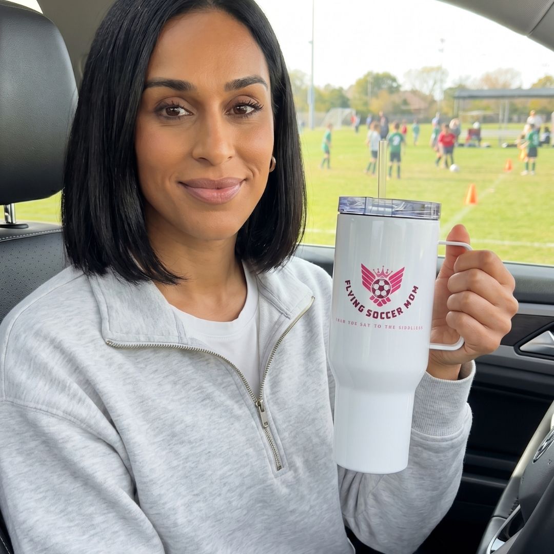 Woman holding a white tumbler with a logo and text, sitting in a car with a soccer field in the background.