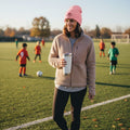 Soccer mom holding a white cup on a soccer field with pink soccer mom beanie