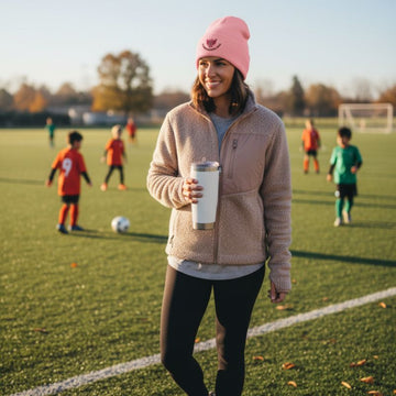 Soccer mom holding a white cup on a soccer field with pink soccer mom beanie