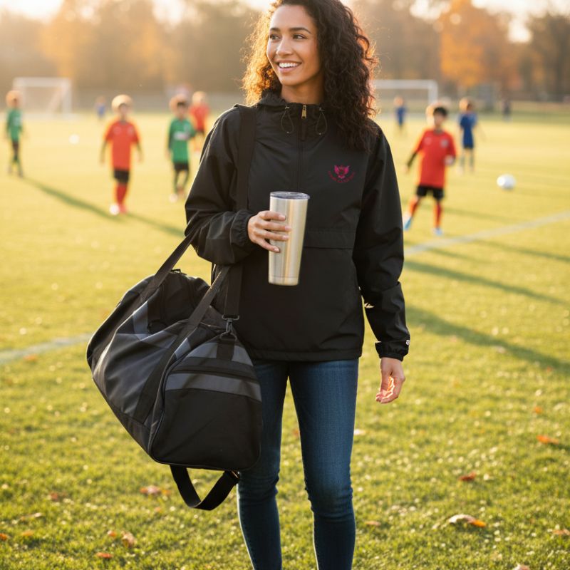 Soccer mom walking on a soccer field holding a duffel bag and a cup, with black soccer mom windbreaker.