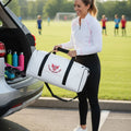 Soccer mom loading a white soccer mom duffel bag with black straps into a car trunk, with a soccer field and players in the background.