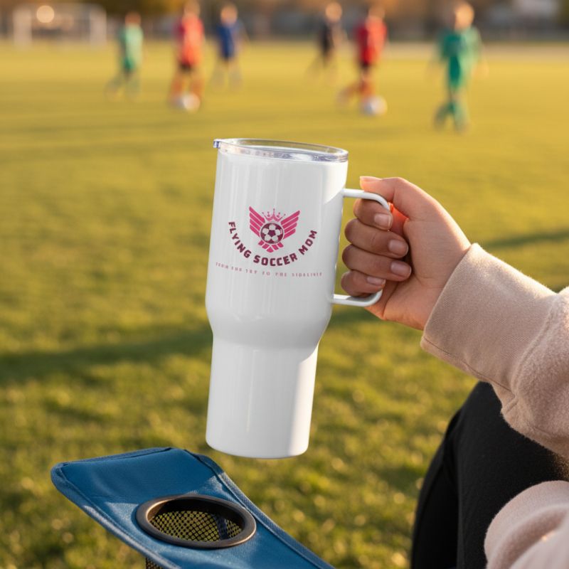 White mug with 'Flying Soccer Mom' logo held by a person on a soccer field